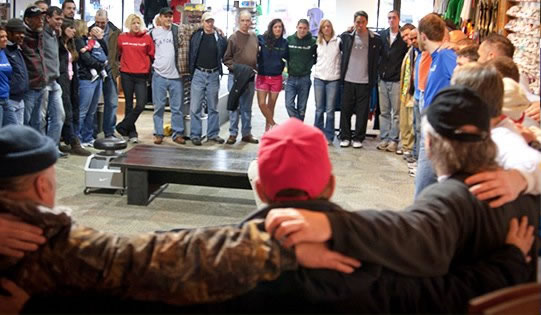 A diverse group of people is standing in a circle with their arms around each other's shoulders. They are indoors, possibly in a community center or meeting space, and are seen from the back. Some individuals are wearing casual clothing, and the atmosphere appears friendly and supportive.