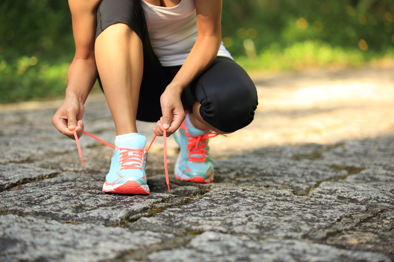 A person wearing a white tank top and black leggings is crouching down to tie the laces of their bright blue and orange running shoes on a cobblestone path surrounded by greenery.