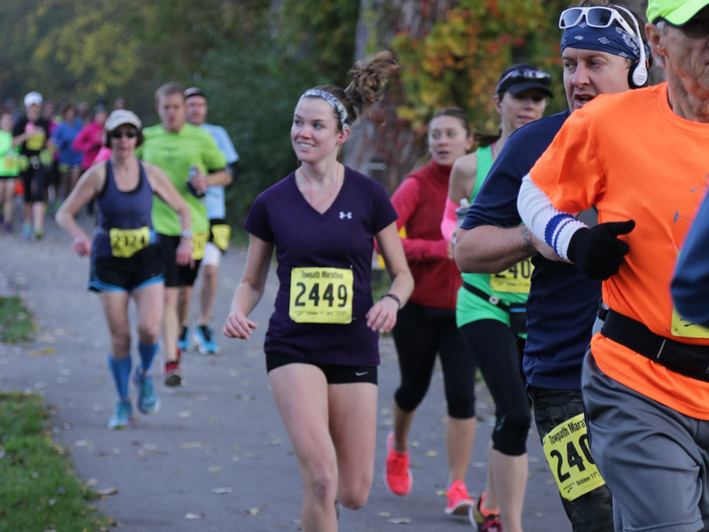 A group of runners participating in a marathon on a paved pathway. The runners are wearing various outfits, bib numbers, and some have headbands or hats. The background features trees, indicating a park-like setting. One runner in a purple shirt smiles while running.