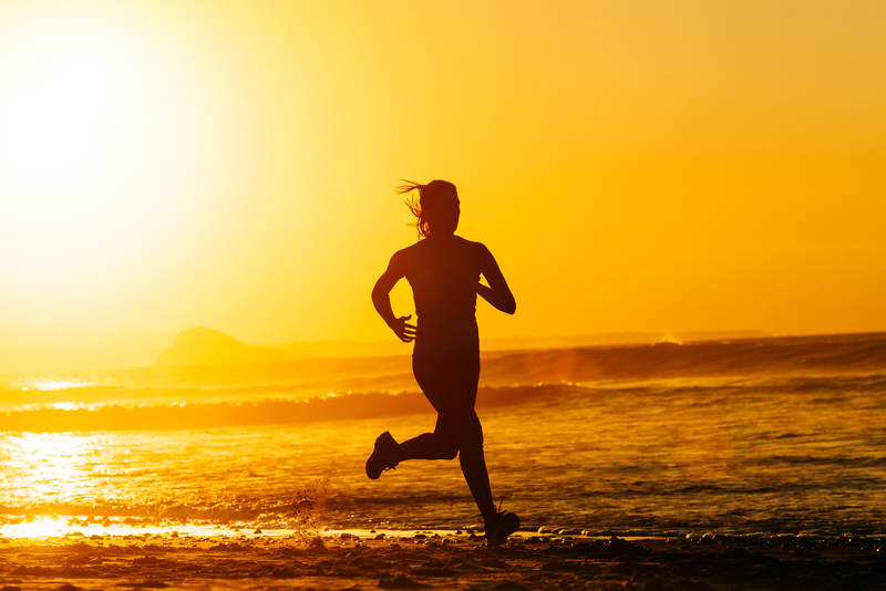 Silhouette of a person running along a beach during sunset, with the sun casting a golden glow over the ocean and sky. The serene scene captures the beauty of nature and the vitality of the runner.