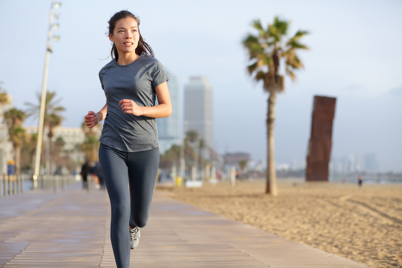 A woman jogs on a wooden pathway by the beach on a clear day. Palm trees and tall buildings are visible in the background, with the ocean to the side. She wears a grey t-shirt and black leggings, looking straight ahead as she runs.