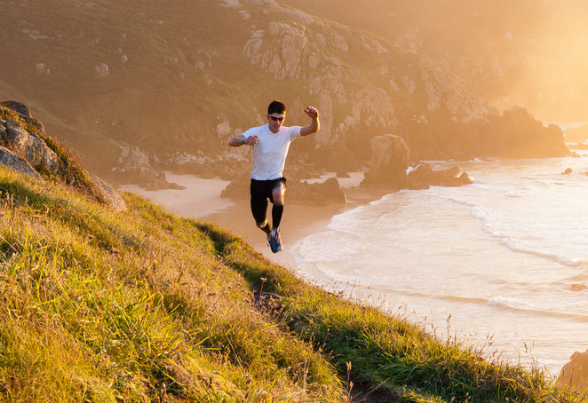 A person wearing a white shirt and black shorts is running energetically down a grassy hill near a coastline at sunset. The ocean waves crash against the rocks below, and the scene is bathed in a warm, golden light.