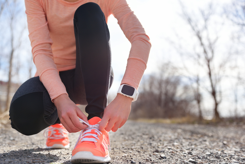 A person in athletic gear crouches to tie the laces on their bright orange running shoes. They are wearing a smartwatch and black leggings. The background features a gravel path and leafless trees, indicating a brisk, likely autumn or winter day.