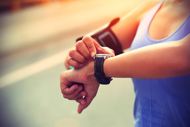 A person wearing a blue tank top checks their smartwatch while standing outdoors. They have an armband on their right arm, likely for holding a phone or music player. The background is slightly blurred, indicating they might be on a run or walk.