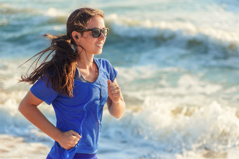 A woman with long hair running along a beach while wearing sunglasses and earphones. She is smiling and dressed in a blue t-shirt, with waves crashing in the background.