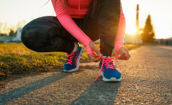 A person with long hair, wearing a bright pink long-sleeve shirt and black camouflage leggings, is crouched down on a paved path at sunset, tying the laces on their blue running shoes with pink laces.