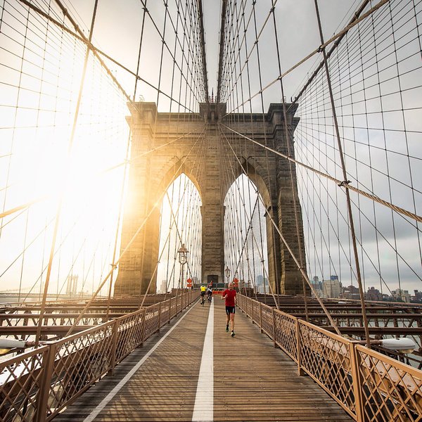 A person in athletic wear jogs across the Brooklyn Bridge during sunrise. The bridge's iconic cables frame the stone arches, and the early morning light creates a warm, glowing effect. The city skyline is faintly visible in the background.