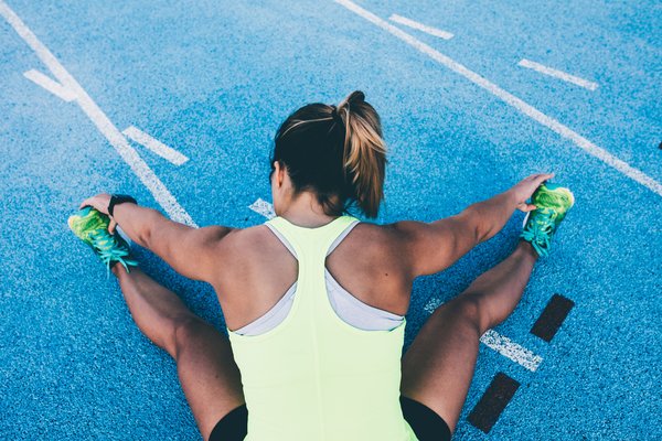 A person with a ponytail and wearing a bright yellow tank top and black shorts is seated on a blue track, stretching their legs apart and reaching to touch their green sneakers with both hands. White lines of the track are visible in the background.