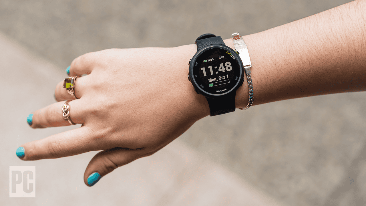 A person with turquoise nail polish and several rings and bracelets on their wrist is wearing a black smartwatch displaying the time as 11:48 and the date as Mon, Oct 7. Equipped with running safety gear, they blend style with functionality. The background is blurred.