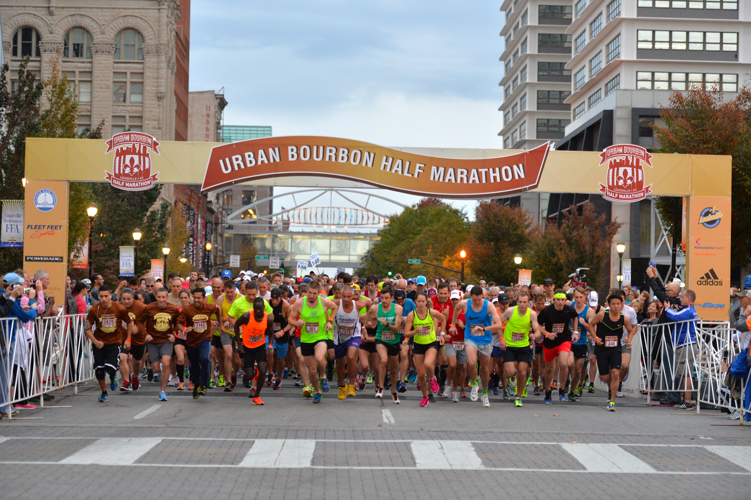 Runners at the starting line of the Urban Bourbon Half Marathon prepare to begin the race. The event banner is visible above, with participants wearing various bright running gear and surrounded by city buildings on a clear day.