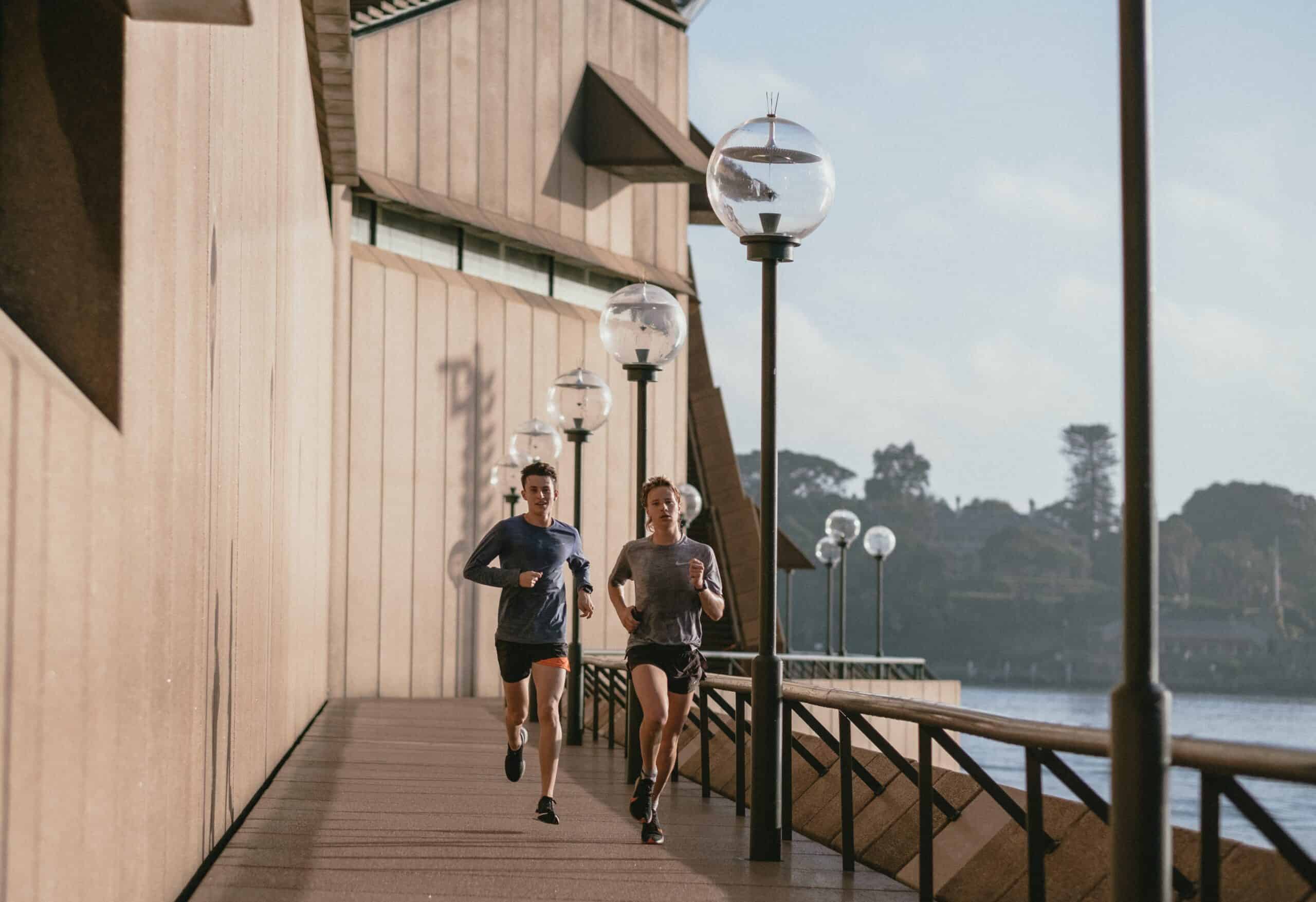 A male and a female runner on a bridge overlooking the water with high running cadence.